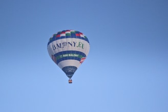 A hot air balloon featuring advertising inscriptions soars against a clear blue sky. The balloon exhibits a variety of colors, including blue, green, and red, creating a vivid contrast with the serene backdrop.