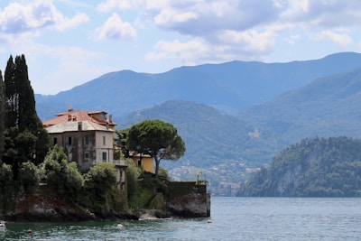 A large villa with a terracotta roof sits by the edge of a lake, surrounded by lush greenery. The villa is framed by tall cypress trees on one side and an expansive view of mountains in the background under a partly cloudy sky.