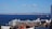 Several cruise ships are docked in a harbor with an ocean and a distant cityscape in the background. The foreground features buildings with red-tiled roofs and various architectural styles.