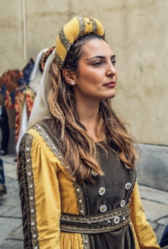 A woman is wearing a detailed medieval-style outfit, featuring a yellow and olive green dress with ornate embroidery and embellishments. She wears a matching yellow headdress. Her expression is serene and poised, suggesting a historical or cultural event. The background is a neutral beige wall.