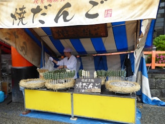 A small food stall featuring a vendor preparing skewers of green and white dango, a Japanese snack, in a market setting. The stall is covered by a blue and white striped tent, with signs in Japanese characters above the display. Straw baskets hold several skewers, and there's a metal grill visible on the counter.