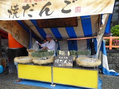 A small food stall featuring a vendor preparing skewers of green and white dango, a Japanese snack, in a market setting. The stall is covered by a blue and white striped tent, with signs in Japanese characters above the display. Straw baskets hold several skewers, and there's a metal grill visible on the counter.