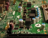 A close-up view of an electronic circuit board featuring various components such as capacitors, resistors, and integrated circuits. The board is mostly green with metallic elements, showing a complex array of electronic architecture.