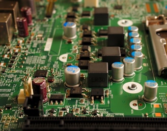 A close-up view of an electronic circuit board featuring various components such as capacitors, resistors, and integrated circuits. The board is mostly green with metallic elements, showing a complex array of electronic architecture.