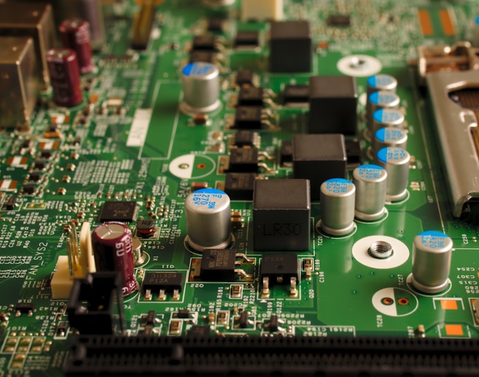 A close-up view of an electronic circuit board featuring various components such as capacitors, resistors, and integrated circuits. The board is mostly green with metallic elements, showing a complex array of electronic architecture.
