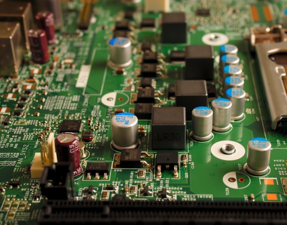 A close-up view of an electronic circuit board featuring various components such as capacitors, resistors, and integrated circuits. The board is mostly green with metallic elements, showing a complex array of electronic architecture.