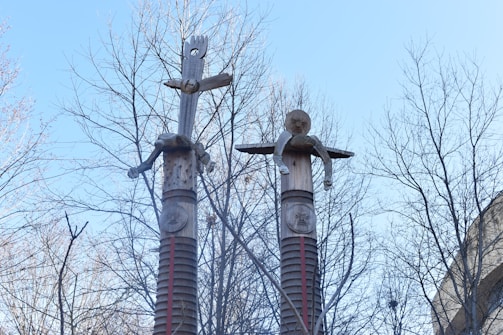 Two tall wooden sculptures resembling totem poles stand against a backdrop of bare tree branches and a clear blue sky. Each sculpture features a carved humanoid figure with distinct facial features. The intricate carvings have a weathered appearance, showcasing details such as arms and circular patterns.