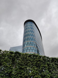 A modern skyscraper with curved blue glass windows rises into a cloudy, overcast sky. The building's sleek design is complemented by a lush green hedge in the foreground.