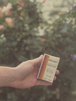 A hand is holding a pack labeled 'RELIEF' with an image of a rhino and text indicating 20 premium filtered hemp cigarettes, 17% CBD, and 0% THC. The background is blurred greenery with soft lighting.
