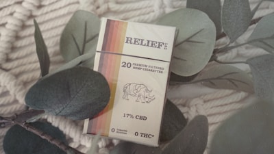 A pack of CBD hemp cigarettes is placed among eucalyptus leaves on a textured, natural fiber background. The packaging features a gradient stripe design and a rhino illustration, with text indicating 17% CBD and 0% THC.