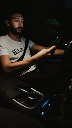 A driver checking his phone while waiting patiently inside a parked vehicle at night.