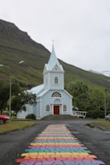 A small, light blue church with a pointed steeple stands in front of a mountainous landscape. In the foreground, a colorful, rainbow-patterned path leads up to the entrance of the church. Lush green trees surround the area, with streetlamps and a parked red car visible to the side. A cloudy sky looms above, adding a serene yet overcast atmosphere.