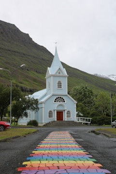 A small, light blue church with a pointed steeple stands in front of a mountainous landscape. In the foreground, a colorful, rainbow-patterned path leads up to the entrance of the church. Lush green trees surround the area, with streetlamps and a parked red car visible to the side. A cloudy sky looms above, adding a serene yet overcast atmosphere.