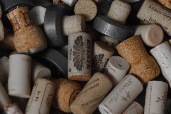 A collection of various wine bottle corks piled together. The corks are made of different materials, including traditional cork and synthetic alternatives. Some have visible branding or logos imprinted on them, with faded text and designs. There are subtle variations in size and shape among the corks.