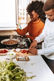 Two individuals are in a kitchen preparing a meal. One person is chopping cherry tomatoes on a wooden cutting board, while the other stirs a pan of food on the stove. Various ingredients are laid out on the counter, including green onions and a roll of sausage. They appear engaged and focused on their cooking activity.