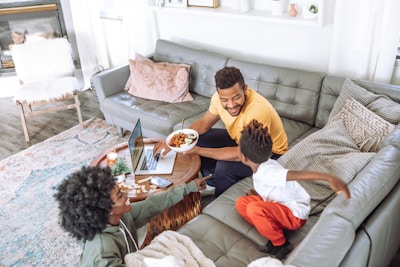 A cozy living room setting with a family enjoying quality time. A man holds a bowl of food and is smiling, interacting with a child who is eagerly reaching for the bowl. A woman with curly hair sits nearby, smiling and engaging in the conversation. The room features a green leather couch, a wooden coffee table, a laptop, and decorative pillows. Soft natural light filters through a window, adding a warm ambiance.