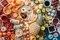 A vibrant assortment of breakfast items and fruits arranged on a table, including eggs, sausage, pancakes, donuts, waffles, and various fruits such as apples, oranges, bananas, and pineapples. There are also beverages like coffee and juice present.