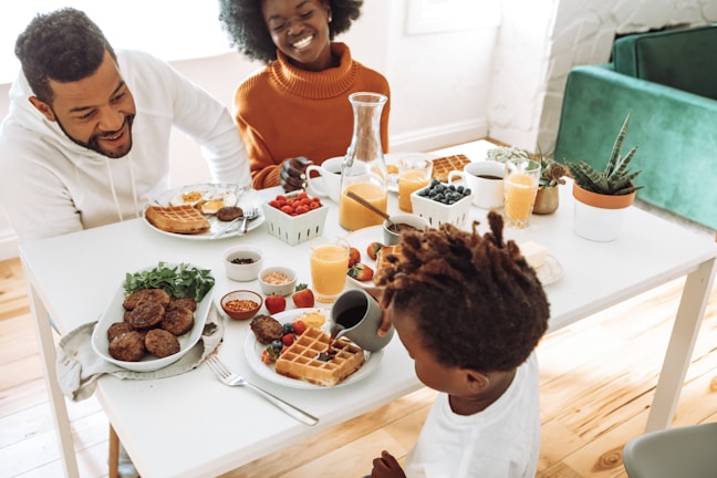 A family enjoys breakfast together at a light-filled table. The table is set with waffles, berries, orange juice, coffee, and a variety of small dishes containing condiments and toppings. Two adults and a child are engaged in a lively conversation, surrounded by plants and a cozy seating area.