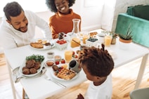 A family enjoys breakfast together at a light-filled table. The table is set with waffles, berries, orange juice, coffee, and a variety of small dishes containing condiments and toppings. Two adults and a child are engaged in a lively conversation, surrounded by plants and a cozy seating area.