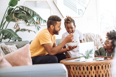 A family scene in a living room with a man in a yellow shirt feeding a child seated on a couch. A woman with a joyful expression is sitting nearby, and a laptop is on a round wooden table. There are large green plants in the background and the room has a cozy, bright atmosphere.
