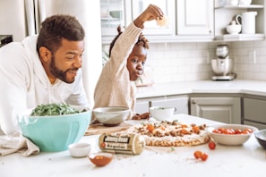 A child sprinkles cheese on a pizza while an adult, possibly a parent, looks on with a smile. The kitchen setting includes a variety of ingredients, such as a bowl of fresh greens and cherry tomatoes, with a clean, modern design. Light streams through a window, illuminating the scene.
