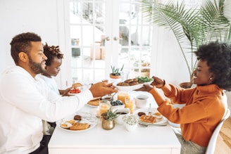 A family enjoys breakfast together at a white dining table. They are passing plates of food, which include waffles, fruit, and juice. The room is bright with natural light coming through large windows with white frames. There are green plants in the background, adding a touch of nature to the setting. The family members are engaged and smiling, creating a warm and friendly atmosphere.