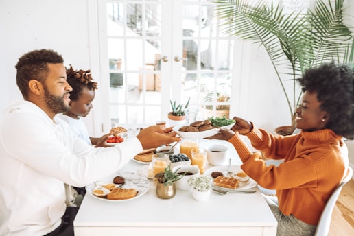 A family enjoys breakfast together at a white dining table. They are passing plates of food, which include waffles, fruit, and juice. The room is bright with natural light coming through large windows with white frames. There are green plants in the background, adding a touch of nature to the setting. The family members are engaged and smiling, creating a warm and friendly atmosphere.