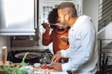 A couple is in a kitchen, engaged in cooking together. One person is tasting food from a spoon offered by the other, creating a joyful and interactive atmosphere.