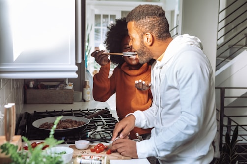 A couple is in a kitchen, engaged in cooking together. One person is tasting food from a spoon offered by the other, creating a joyful and interactive atmosphere.