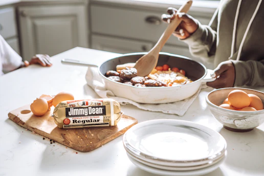 A person is cooking a meal in a kitchen, stirring a frying pan with a wooden spoon. The pan contains sausages and vegetables. Next to the pan is a chopping board with a roll of Jimmy Dean sausage and several eggs. A bowl filled with more eggs and a stack of plates are also visible on the counter.