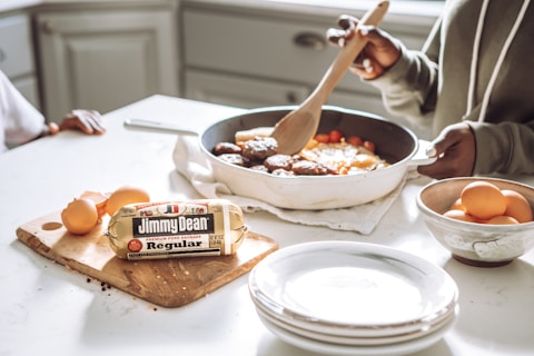 A person is cooking a meal in a kitchen, stirring a frying pan with a wooden spoon. The pan contains sausages and vegetables. Next to the pan is a chopping board with a roll of Jimmy Dean sausage and several eggs. A bowl filled with more eggs and a stack of plates are also visible on the counter.