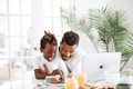 A father and his young son share a joyful moment at a breakfast table. The father has his arm around the son, who is laughing and engaging with the food. The table is set with bowls of fruit, glasses of juice, and a plate of waffles. A laptop is open in front of them, and there is a large, lush plant in the background, creating a bright and cozy atmosphere.