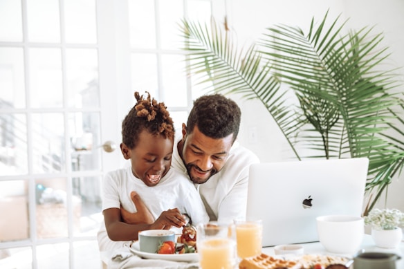 A father and his young son share a joyful moment at a breakfast table. The father has his arm around the son, who is laughing and engaging with the food. The table is set with bowls of fruit, glasses of juice, and a plate of waffles. A laptop is open in front of them, and there is a large, lush plant in the background, creating a bright and cozy atmosphere.