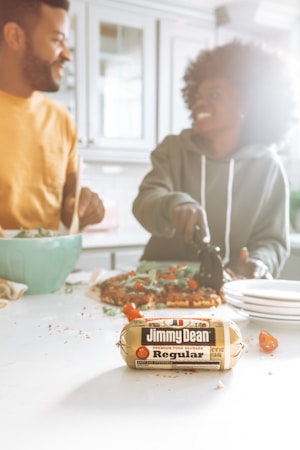 A kitchen scene featuring two people engaging in meal preparation. A man wearing a mustard yellow shirt and a woman in a dark green hoodie are smiling and enjoying their time together. A package of Jimmy Dean regular pork sausage is prominently placed on the kitchen counter in the foreground. There is a salad in a bowl and a pizza being prepared in the background, along with scattered vegetables.