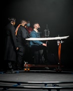 A group of individuals wearing face masks is gathered around a camera setup in a dimly lit environment. One person is seated on a dolly, focusing on operating the camera, which is mounted on a track. The scene has a professional and focused atmosphere, likely depicting a film or video production setting.