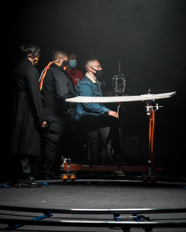 A group of individuals wearing face masks is gathered around a camera setup in a dimly lit environment. One person is seated on a dolly, focusing on operating the camera, which is mounted on a track. The scene has a professional and focused atmosphere, likely depicting a film or video production setting.