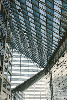 A large glass atrium with an intricate metal framework, showcasing the modern architecture of a building. The structure features a series of triangular glass panels supported by steel beams. Several large, industrial ceiling fans are visible, and the surrounding walls are composed of glass windows with metal casings.