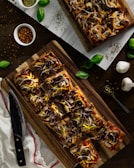 Rectangular pizzas topped with olives, onions, mushrooms, and cheese are showcased on wooden boards. Fresh basil leaves are scattered around, along with garlic bulbs, a bowl of pepper flakes, and dried herbs.