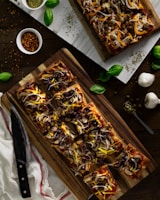 Rectangular pizzas topped with olives, onions, mushrooms, and cheese are showcased on wooden boards. Fresh basil leaves are scattered around, along with garlic bulbs, a bowl of pepper flakes, and dried herbs.