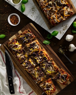Rectangular pizzas topped with olives, onions, mushrooms, and cheese are showcased on wooden boards. Fresh basil leaves are scattered around, along with garlic bulbs, a bowl of pepper flakes, and dried herbs.