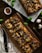 Rectangular pizzas topped with olives, onions, mushrooms, and cheese are showcased on wooden boards. Fresh basil leaves are scattered around, along with garlic bulbs, a bowl of pepper flakes, and dried herbs.