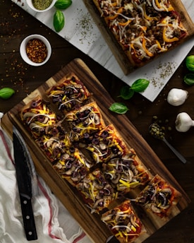 Rectangular pizzas topped with olives, onions, mushrooms, and cheese are showcased on wooden boards. Fresh basil leaves are scattered around, along with garlic bulbs, a bowl of pepper flakes, and dried herbs.