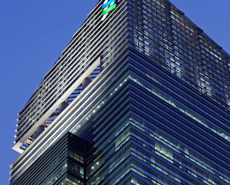 A modern skyscraper with sleek glass panels and a prominent logo at the top. The architecture features a mix of vertical and horizontal lines, creating a striking geometric design against the evening sky.