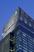 A modern skyscraper with sleek glass panels and a prominent logo at the top. The architecture features a mix of vertical and horizontal lines, creating a striking geometric design against the evening sky.