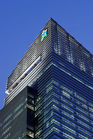 A modern skyscraper with sleek glass panels and a prominent logo at the top. The architecture features a mix of vertical and horizontal lines, creating a striking geometric design against the evening sky.