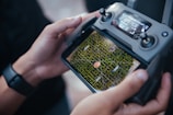 A person is holding a drone controller with a screen displaying an aerial view of a green maze. The controller has various buttons and a digital display showing GPS mode and other data. The person's hands are visible, and they are wearing a smartwatch.