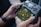 A person is holding a drone controller with a screen displaying an aerial view of a green maze. The controller has various buttons and a digital display showing GPS mode and other data. The person's hands are visible, and they are wearing a smartwatch.