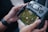 A person is holding a drone controller with a screen displaying an aerial view of a green maze. The controller has various buttons and a digital display showing GPS mode and other data. The person's hands are visible, and they are wearing a smartwatch.