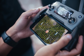 A person is holding a drone controller with a screen displaying an aerial view of a green maze. The controller has various buttons and a digital display showing GPS mode and other data. The person's hands are visible, and they are wearing a smartwatch.