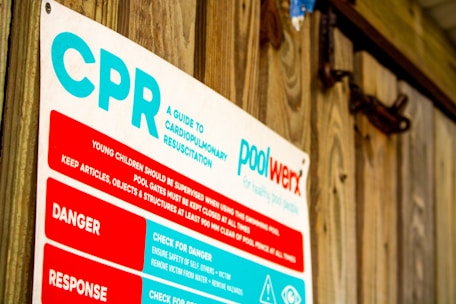 A sign detailing cardiopulmonary resuscitation (CPR) instructions, positioned against a wooden wall. The text includes warnings and response tips and highlights the importance of supervision around pools.